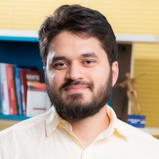 Caio César Lopes sorrindo, de barba e camisa clara, em frente a uma estante de livros