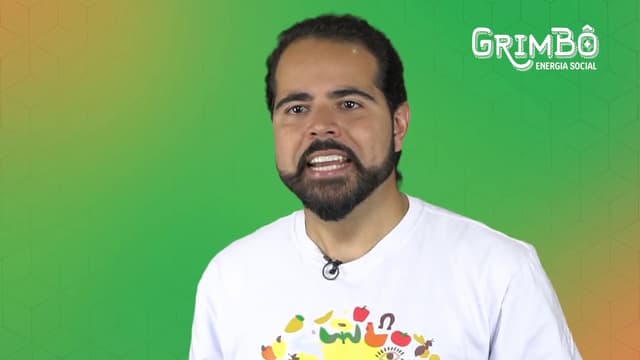Apresentador de camiseta branca falando para a câmera sobre fundo verde e laranja, com a marca Grimbô